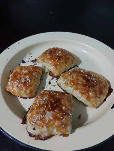 A top-down shot shows four golden-brown pastries sprinkled with white sesame seeds, arranged on a light yellow plate. The pastries appear to be some type of baked goods, possibly savory rolls or turnovers, with flaky, slightly browned crusts. Crumbs are scattered on the plate and around the pastries, indicating they have been handled or are being served. The plate itself has a slightly worn, enamel appearance, suggesting it might be a common household item. The background is dark and out of focus, creating a contrast that highlights the food. The lighting suggests an indoor setting, and given the pastries appear freshly baked, it's likely a mealtime or snack time. There is no visible text in the image. The scene is simple, focusing entirely on the appetizing presentation of the pastries.