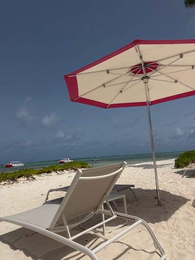 This is a medium-angle, eye-level shot of a tranquil beach scene in Higüey, Dominican Republic. The foreground features two white and gray beach chairs on a sandy shore. One chair is closer to the viewer and is partially reclined. In the mid-ground, a large, octagonal umbrella with a white canopy and a pink border provides shade. The umbrella pole is planted in the sand. Beyond the umbrella, the calm, turquoise ocean stretches to the horizon. Two white boats with red accents are visible on the water, along with a thin line of lush green vegetation along the shoreline. The sky is a deep blue with scattered white clouds. The overall impression is one of peaceful relaxation and a clear, sunny day.