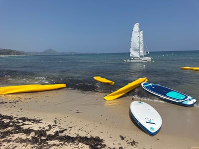 This media file depicts a scenic beach scene in Castiadas, Italy, under a clear, sunny sky. The foreground features a sandy beach littered with seaweed. Several recreational watercraft are present: two yellow kayaks are partially on the sand and partially in the shallow water, one yellow kayak is floating further out, and two stand-up paddleboards, one white with blue accents and the other blue with white and turquoise accents, are on the sand near the water's edge. In the background, a white catamaran with two sails is sailing on the blue ocean. The water is calm, with small waves gently lapping the shore. Distant hills and a mountainous coastline are visible in the far background under a vast blue sky, suggesting a warm, pleasant day. No people are visible in the image, and the overall mood is serene and tranquil. Visible text on the paddleboards includes "TAHE" and "SUP-YAK".