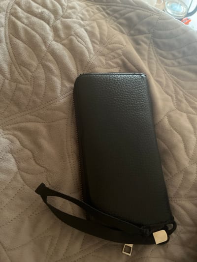 The image shows a black, pebbled leather clutch or wallet resting on a textured, light brown blanket. The clutch has a wrist strap made of black fabric that is attached to a metallic silver buckle. The blanket has a quilted pattern of wavy lines. In the upper left corner, out of focus, there are some glass objects, possibly jars or containers. The lighting suggests it is daytime, and the overall scene appears to be indoors, perhaps on a bed or couch. There is no visible text in the image. The setting is likely a home environment in Sergeevka, Russia. The image focuses on the object and its texture, with no people or specific activities depicted.