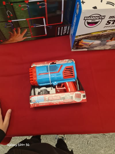 The image, captured in Keizer, Oregon, United States, showcases a collection of retail items laid out on a red surface. In the foreground, a blue and red bubble gun is prominently displayed within its packaging. The packaging indicates that the gun has 36 holes, comes with 80ml of non-toxic bubble solution, and requires 4 AA batteries (not included). A QR code is visible on the packaging, suggesting a link to a product video. To the left of the bubble gun, a person's hand and arm are visible, seemingly reaching towards another item. To the right, a portion of a large cardboard box is visible, featuring the "BIGMOUTH INC." logo and an illustration of people enjoying a pool. The background is partially obscured by these items, but appears to be a retail environment. The overall scene is brightly lit, with the red surface dominating the composition.