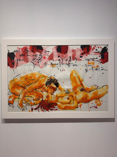 This is a framed watercolor painting of two nude figures in an abstract style, mounted on a white wall. The figures are rendered in warm orange and yellow tones with black ink outlines. They are positioned in a dynamic, intertwined pose, with one figure appearing to be upside down and supported by the other. The background is predominantly white, with splashes of red and black ink, as well as some illegible black calligraphic text along the left side of the painting. The style is expressive and gestural, with visible brushstrokes and drips of watercolor. The lighting suggests an indoor setting, likely a gallery or exhibition space. There is no indication of time of day, weather, or specific location within the artwork itself, other than the provided context of La Cuesta, Spain. The overall mood is artistic and somewhat sensual, with the figures appearing relaxed or in a moment of contemplation or embrace.