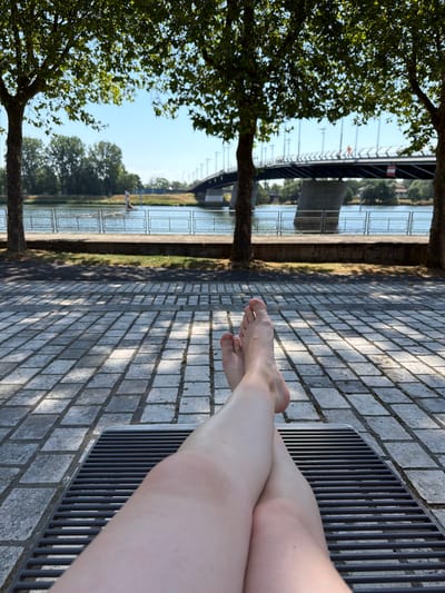 The image depicts a pair of legs and feet crossed and resting on a metal grate, suggesting a person is reclining outdoors. The foreground features a cobblestone pathway with dappled sunlight, indicating an open, paved area. Beyond the pathway, a body of water, likely a river, is visible, bordered by a railing. In the middle ground, trees line the riverbank, and a bridge spans the water in the background. The sky is clear and blue. The location is identified as Charnay-lès-Mâcon, France. The scene conveys a sense of leisure and relaxation in an urban park or waterfront setting.