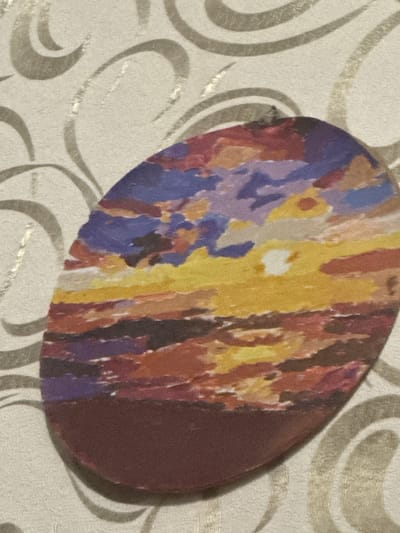 An oval-shaped artistic depiction of a sunset is the central subject of the image. The artwork features a vibrant, impressionistic rendering of the sky with bold brushstrokes of yellow, orange, red, purple, and blue, suggesting clouds and the setting sun. The lower portion of the oval is a solid dark reddish-brown color, likely representing land or water. The artwork is placed on a textured surface with a light-colored background adorned with a swirling, grey, decorative pattern. The image is a close-up view, with no context provided regarding its specific location in the city of Гожули, Ukraine, or any surrounding environment.