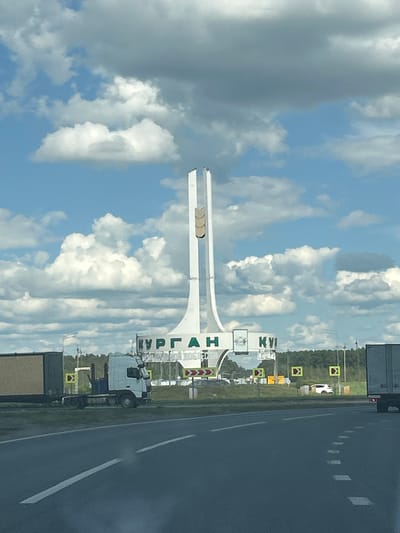 The image shows a monument and sign in the city of Kurgan, Russia, on a cloudy day. The monument is tall and white, with a distinctive shape. The sign behind it has green lettering on a white background, spelling out "КУРГАН" (KURGAN) and "КУ" (KU). In the foreground, there is a road with white lane markings. A white truck with a trailer is parked on the side of the road to the left. To the right, a white car is visible in the distance. There are several traffic signs with yellow and red arrows indicating directions. The environment appears to be a highway or a road leading into the city.