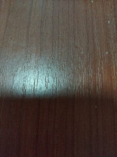 The image displays a close-up, overhead view of a dark brown, textured surface. The surface exhibits a prominent vertical grain pattern, consistent with wood or wood-grain laminate. A bright, elongated glare, appearing whitish-blue, is centered in the upper-middle portion of the image, indicating a direct light source reflecting off the surface. Scattered across the surface, particularly visible in the upper right quadrant, are numerous small, light-colored specks, consistent with dust or minor surface imperfections. The lower portion of the image is darker, suggesting less direct illumination or a slight shadow. No people, specific objects beyond the surface material, actions, or interactions are visible. The specific location within Logatec, Slovenia, cannot be determined from the visual information provided.