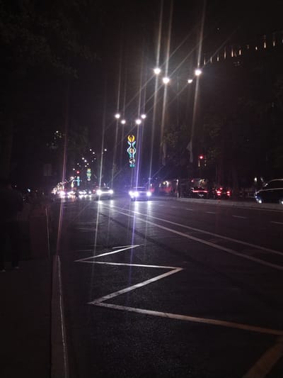 The image depicts a street scene at night, possibly in Kashgar Mahalla, Uzbekistan, with several cars driving on a wet asphalt road. The street is illuminated by overhead streetlights, which create strong light streaks due to the camera's aperture. Decorative colorful lights are strung along the street, adding to the ambiance. On the left side of the road, a sidewalk is visible with a few people standing. Trees line the sides of the street, their dark silhouettes contrasting with the artificial lights. The road surface is speckled with white lines, indicating lanes and possibly a pedestrian crossing. The overall impression is of a lively urban environment at night, with traffic and decorative lighting.