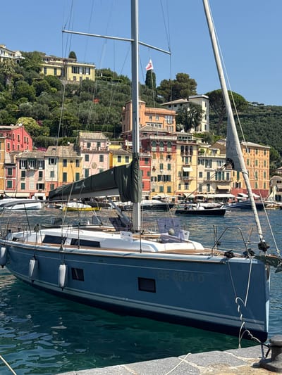 The image depicts a serene harbor scene in Portofino, Italy, on a clear, sunny day. In the foreground, a blue sailboat is moored, its hull displaying the registration "GE 8624 D". The sailboat's mast and rigging are prominent, with a furled sail and a small flag at the top. The water is calm and a deep teal color, with reflections of the surrounding buildings.
The background is dominated by the iconic colorful buildings of Portofino, cascading down a lush, green hillside. The buildings are painted in vibrant shades of yellow, pink, orange, and red, with traditional Italian architectural features like arched windows and balconies. The hillside is densely covered with trees and vegetation, suggesting a Mediterranean climate. Other boats, including motorboats and smaller vessels, are visible in the harbor, further out from the main sailboat. A stone quay is visible in the lower right corner, with a metal bollard for mooring. The sky is a bright, cloudless blue. No people are clearly visible in the image. The overall impression is one of a picturesque and tranquil coastal town.