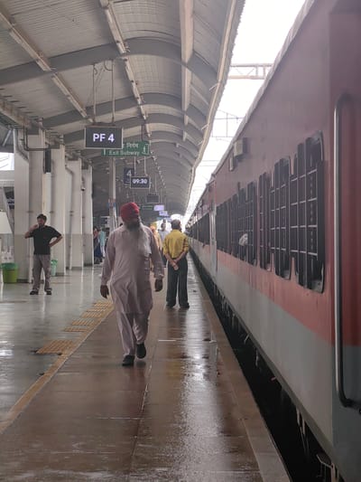 The image depicts a railway platform at the Bhopal Junction railway station in Bhopal, India. A red and white train is parked on the right side of the frame. On the platform, a man wearing a light-colored kurta and a red turban walks towards the foreground. To his left, a man in a black t-shirt and grey pants stands and drinks from a glass. Further down the platform, another man in a yellow shirt and dark pants stands with his back to the camera. Several other people are visible in the background, some walking and some standing. Overhead, a canopy provides shelter, with signs indicating "PF 4" and "Exit Subway." The platform is wet, suggesting it may have rained recently. The overall scene captures a typical moment at an Indian railway station.