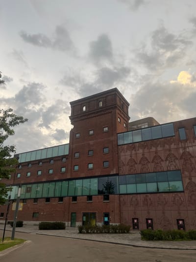 The image shows the exterior of a brick building in Enschede, Netherlands. The building features a tall, square tower with arched windows and a series of horizontal glass windows along its facade. The building's lower section is decorated with a relief of figures on scooters. The sky above is cloudy with some patches of light. A paved walkway and some greenery are visible in front of the building.