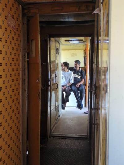 The image captures the interior of a train compartment, viewed from a narrow doorway. The left side of the frame is dominated by a wall with a yellow and brown patterned wallpaper. A pink and white informational card is affixed to this wall. To the right of the wallpapered section, an open, orange-colored door reveals a hallway or passage.

Further into the compartment, two men are seated side-by-side. The man on the left wears a white t-shirt with "DIESEL" printed on it and dark pants. He has dark, curly hair and is looking to his left. The man on the right wears a black polo shirt with a striped collar and dark pants. He has short, dark hair and a mustache and is looking towards the left, away from the viewer. Both men are seated on a bench that appears to be part of the train's seating.

The background of the compartment shows more of the train's interior, including metal walls and doors. The floor is covered with a textured, dark material. The overall impression is of a functional, somewhat aged train carriage. The city of Bithooda, India, is a small village in Rajasthan, known for its temples. This image is consistent with the interior of an Indian train.