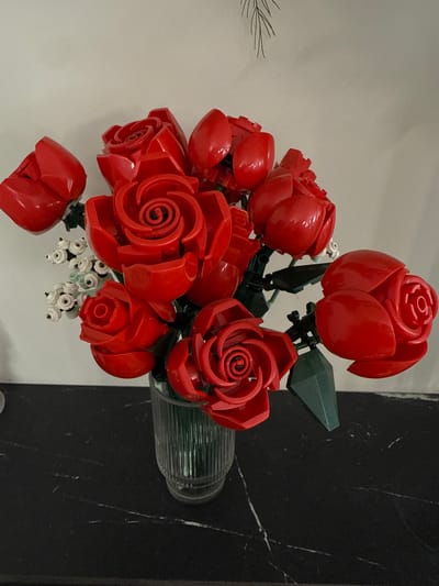 A bouquet of red roses, constructed from Lego bricks, is displayed in a ribbed glass vase. The vase sits on a black marble countertop against a plain white wall. The roses are a mix of open blooms and buds, with green Lego leaves and stems supporting them. To the left of the roses, a small cluster of white Lego flowers with black centers is visible. A delicate line drawing of a pine branch hangs on the wall above the bouquet. The scene is lit from the front, casting subtle shadows. The location is in the city of Khokhryaki, Russia.
