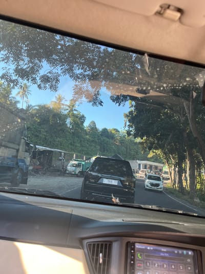 The image shows a view from inside a vehicle, looking forward through the windshield. The windshield is partially obscured by streaks and smudges. Outside, a road scene is visible on a sunny day. Several vehicles are present, including a large blue truck on the left, a black SUV with license plate "L 1948 FF" in the foreground, and white cars further down the road. There are also some smaller trucks or vans. To the left of the road, there appears to be a small, informal settlement or workshop area with a covered structure and various items stored. The background consists of a lush green hillside covered in trees and vegetation, with some palm trees visible. The sky is clear and blue. The car interior shows part of the dashboard and a display screen. The scene appears to be a roadside in a tropical or subtropical environment, consistent with the location in Pekutatan, Indonesia, which is known for its natural landscapes and rural settings. There are no clearly identifiable individuals engaged in specific actions.