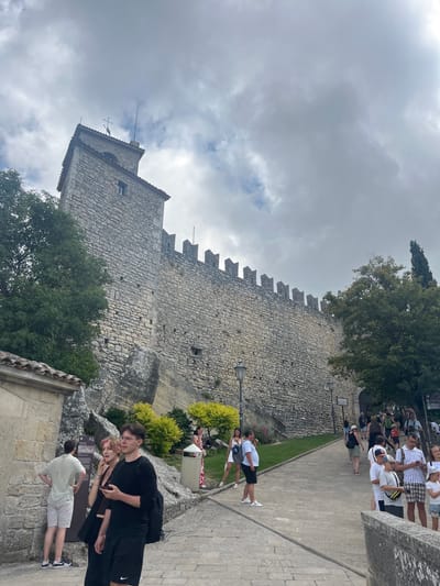 The image shows a view of the Guaita Tower, the oldest of the three towers of San Marino, located in the city of San Marino, San Marino. The tower is made of stone and has crenellated battlements. A stone wall extends from the tower, also featuring battlements. In the foreground, a group of tourists are walking on a paved path. Some are looking at their phones, while others are observing the scenery. The path is lined with trees and some green foliage. The sky is overcast with clouds.