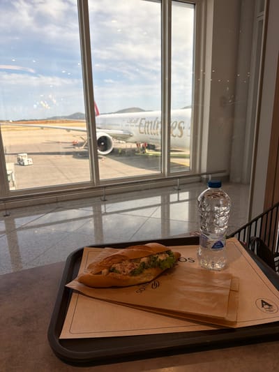 The image shows a tray on a table in what appears to be a dining area, possibly an airport lounge, in Spata-Loutsa Municipal Unit, Greece. The tray holds a sandwich, a bottle of water, and a brown paper wrapping. Through a large window, an Emirates airplane is visible on the tarmac. The sky is partly cloudy.