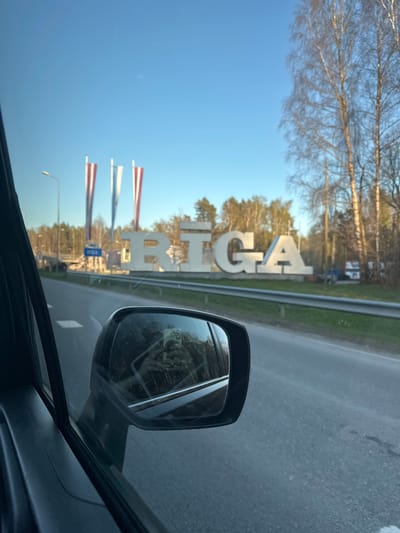 The image depicts a view from inside a car, likely moving, towards the Riga sign near Spilve, Latvia. The large white letters "RĪGA" are visible on the side of the road with Latvian flags flying in the background. The car's side mirror is in the foreground, reflecting the surrounding environment. The environment contains trees, a highway, and a clear blue sky.