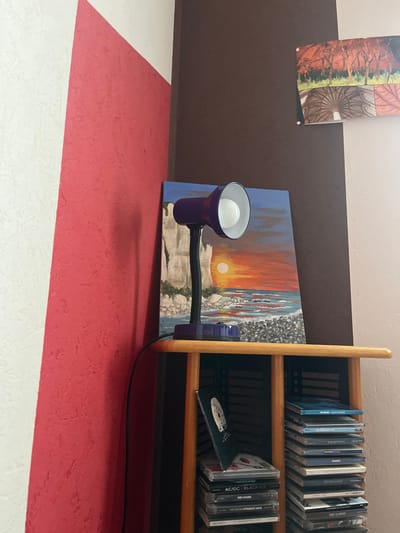 The scene is situated in a room in Ancenis, France. The image captures an indoor corner where two walls meet. The left wall features a textured white surface partially covered by a diagonal, textured red paint stripe. The right wall is divided vertically, with a darker brown textured section on the left and a lighter beige or off-white textured section on the right.
A light-colored wooden shelf unit is positioned in the corner. On the top surface of this unit, a purple desk lamp with a flexible black neck and a white interior reflector is present, illuminating a visible light bulb. To the right and behind the lamp, an unframed rectangular painting depicts a sunset over a body of water with prominent white cliffs on the left, a bright orange sun setting over the horizon, and a foreground of grey pebbles or rocks.
Higher on the brown section of the right wall, a horizontally oriented framed or mounted picture is displayed. This picture appears to be a diptych or collage, with the left half showing autumn trees with red foliage and the right half displaying an architectural or geometric pattern with circular elements in earth tones.
On the lower shelf of the wooden unit, two stacks of compact disc (CD) cases are visible. The left stack includes a partially obscured case with a white circular graphic on a dark background, and cases labeled "AC/DC Black Ice" and "The Doors Strange Days." The right stack is taller, with several CD cases displaying blue or light-colored spines, including "Joe Satriani - Crystal Planet" at the top. A black power cord for the desk lamp extends downwards along the left wall. No individuals are present within the frame.