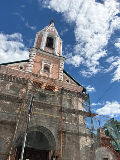 This image shows the bell tower and part of the main building of a church in Pereslavl-Zalessky, Russia, under renovation. The building's facade is made of pink brick with white decorative elements. The bell tower has a white spire with a golden cross. The main building is partially covered with scaffolding and a green mesh net, suggesting ongoing construction or repair work. Wooden beams and planks are visible through the netting. A small arched window is visible on the pink brick section of the building. The sky is bright blue with scattered white clouds. The sun is shining, indicating it is daytime. The overall scene is of historical architecture undergoing preservation. No people are visible in the image. There is no visible text.