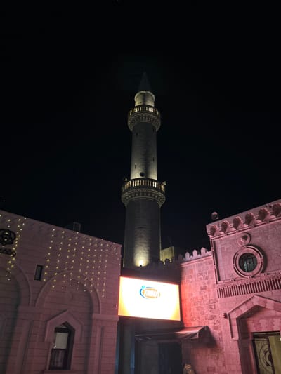 This image presents a nighttime street-level view in Amman, Jordan. Centrally positioned and occupying the majority of the vertical frame is a slender stone minaret, illuminated from its base and balconies by upward-facing warm yellow lights, culminating in a dark conical spire against a black sky. Below the minaret, a large rectangular LED display screen exhibits a bright 'JORINOS' logo, rendered in blue and orange. To the immediate left, a light-colored building constructed of block-like stone features multiple arched window openings and an upper facade decorated with a uniform grid of small, amber-colored string lights, alongside a circular dark emblem. To the right, a separate stone building, distinguished by a reddish ambient light, includes a crenellated roofline, a circular window with an ornate frame, and a deeply recessed arched entrance. A single individual, clad in light-colored garments, is partially visible at the base of the LED screen, within the shadow of an entryway. The scene is illuminated by multiple artificial light sources.