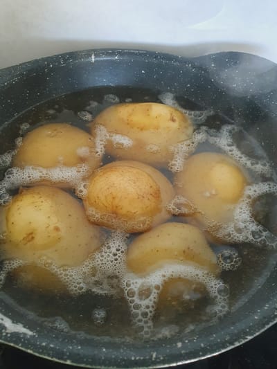 A close-up, overhead shot shows several whole, unpeeled potatoes boiling in a dark gray pot. The potatoes are a light brown to yellow color, with some visible blemishes and lines on their skins. The water is cloudy and filled with small bubbles, indicating that it is actively boiling. The rim of the pot is visible, showing a speckled pattern with some wear. The background is a blurred, light-colored surface. The image appears to be taken in a kitchen setting.