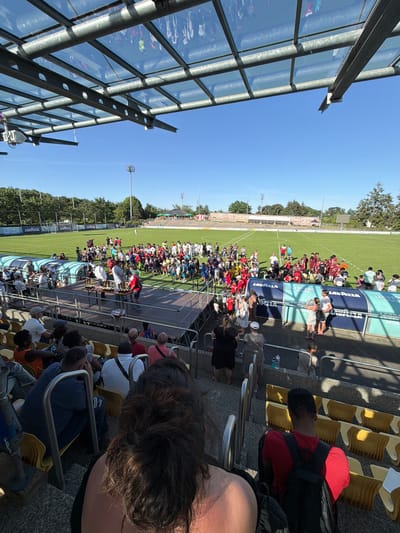 The image shows a crowd of people gathered on a soccer field in Fentange, Luxembourg, likely for an award ceremony. They are standing between the sidelines and a sheltered dugout. People are dressed in different colors, including many in athletic wear and some in red or white jerseys. Some are holding trophies on a small platform. Spectators sit in tiered seating to one side, and a glass and metal structure is visible on the other side. A tall row of trees lines the edge of the field. The sky is clear and blue.