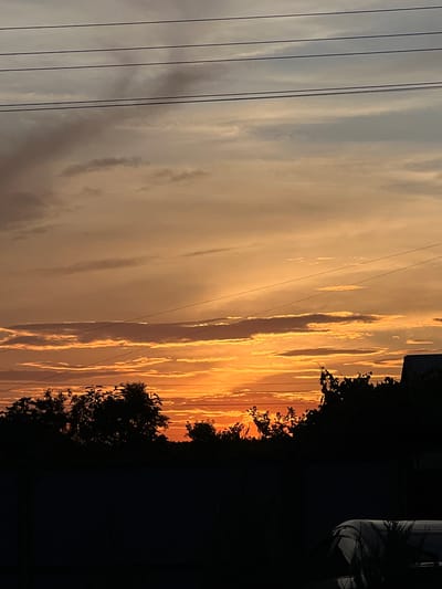 This media file shows a dramatic sunset over Kulishi, Ukraine. The sky is a vivid spectacle of orange and yellow hues, with streaks of clouds catching the last rays of sunlight. The scene is framed by silhouetted trees on the right side, adding a sense of depth and contrast to the fiery sky. Several power lines stretch across the left side of the image, interrupting the smooth gradient of the sky and creating a sense of civilization within the natural landscape. The overall impression is one of natural beauty and the serene transition from day to night. The time of day is clearly sunset, indicated by the warm, saturated colors in the sky. While no people are visible, the presence of power lines suggests a populated area, though the natural elements of the sky and trees dominate the frame. The mood is tranquil and awe-inspiring, evoking the beauty of a Ukrainian sunset. There is no visible text in the image.