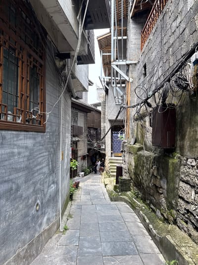 An alleyway in Fenghuang, China, with a stone-paved path between buildings. One side features a weathered stone wall covered in moss, while the opposite side shows gray-tiled buildings with windows, some with ornate wooden lattice frames. Utility cables tangle overhead, interspersed with hanging objects, possibly clothing. People are seen at the far end of the alley. The image is oriented with the alley running vertically.