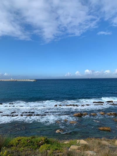 This image captures a coastal landscape in Ksar El Majaz, Morocco, viewed from an elevated position. The scene features a clear blue sky occupying the upper third, adorned with scattered white cumulus clouds. The horizon line is distinct.
The middle ground is dominated by the deep blue-green waters of the sea. Closer to the foreground, white foamy waves crash against and flow around numerous dark brown and reddish-brown rocky outcrops that extend into the water. The water in shallower areas appears translucent, revealing parts of the submerged rock formations.
In the far left-midground, a substantial grey concrete breakwater or pier extends horizontally into the sea, terminating with a small, light-colored lighthouse or navigational beacon. Behind the breakwater, indistinct industrial or port structures are visible on the distant coastline. On the far right horizon, several large vessels are faintly visible.
The immediate foreground consists of a rugged, rocky terrain sparsely covered with low-lying green and yellow-brown coastal vegetation, including succulent-like plants. No individuals, specific actions, or direct interactions are visible within the frame.