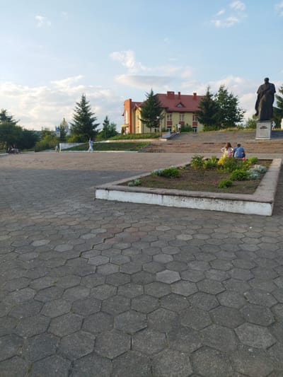 In Novyi Rozdil, Ukraine, a paved plaza made of hexagonal grey stones is in the foreground. A raised flower bed with green plants is in the center of the plaza. Beyond the plaza, a wide set of stairs leads up to a building with a red roof and yellow walls. A dark bronze statue of a man in a robe stands to the right of the stairs, on a pedestal. In the background, a golden-domed church is visible, surrounded by trees. Several people are scattered throughout the scene: a child on a bicycle rides near the stairs, a woman walks away from the camera towards the church, and a couple sits together in the flower bed. Other figures are present on the stairs and in the distance. The sky above is blue with scattered white clouds.