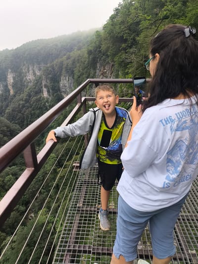 In the city of Satsiskvilo, Georgia, a boy with light brown hair and a green and blue t-shirt, black shorts, and gray sneakers stands on a metal observation deck. He is sticking his tongue out at the camera. Next to him, a woman with dark hair, glasses, and a white t-shirt with blue lettering stands with her back to the camera, holding a phone to take a picture of the boy. The observation deck overlooks a deep canyon filled with lush green trees. The sky is overcast and foggy.