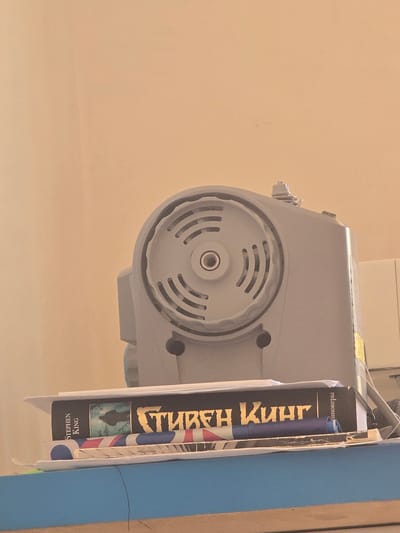 The image shows a close-up of a sewing machine, a book, and other items on a shelf. The sewing machine is gray and has a circular vent on the top. The book is black with gold lettering and appears to be in Russian. The spine of the book has the name "STEPHEN KING" printed vertically. A thin black wire is visible in the foreground. The background is a plain, light-colored wall. The scene is indoors, on a shelf, likely in a domestic setting. There is no information to determine the specific location in the city of Avtury, Russia.