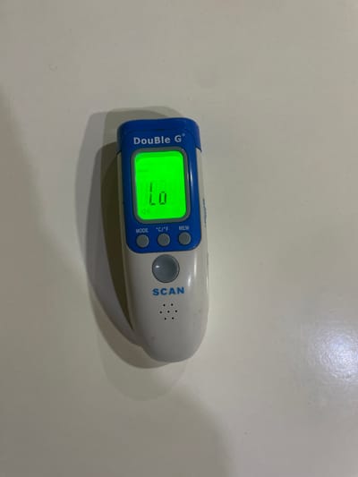 A "DouBLe G" brand infrared thermometer, white with a blue top section, rests on a light-colored, flat surface in Nibo, Nigeria. The device is oriented vertically, with its display screen illuminated in green. The screen shows "Body" in the top left corner, "Lo" prominently in the center, and a speaker icon below the "Lo" reading. Three small gray circular buttons labeled "MODE", "°C/°F", and "MEM" are located below the screen. A larger central gray circular button is positioned further down, above the word "SCAN" printed in blue. Seven small circular perforations, likely a speaker grille, are visible below the "SCAN" text. The thermometer exhibits minor surface wear and scuffs. Light sources illuminate the scene from the upper left, casting a soft shadow of the device onto the background surface. No other objects or individuals are present in the immediate vicinity.