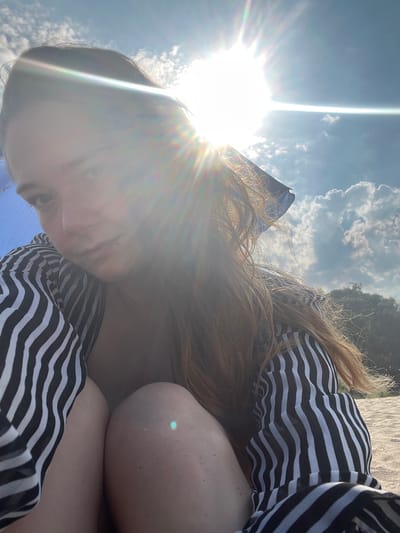 The image is a selfie taken outdoors, with the sun shining brightly and casting lens flares. The subject is a person lying down, with their face partially obscured by the sun. They are wearing a black and white striped top. The background shows a bright blue sky with scattered clouds, suggesting a clear day. There are also hints of greenery and sand, indicating an outdoor setting like a beach or park. The strong sunlight and the angle of the shot suggest it is daytime, likely late morning or afternoon. The person's pose and the relaxed atmosphere hinted at by the setting suggest a leisure activity. No visible text is present in the image.