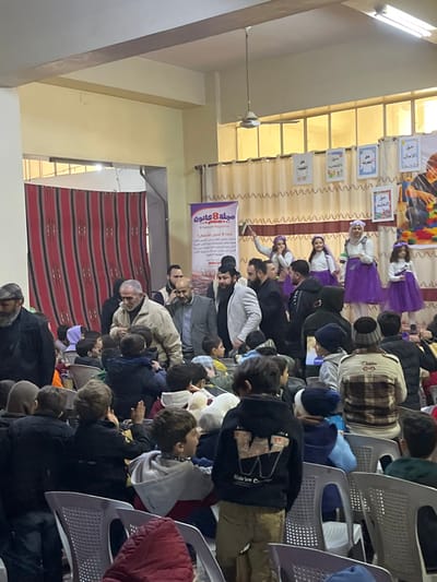 This image depicts an indoor event in Kafr Batna, Syria, featuring a performance for an audience. In the background, a raised stage area is visible with five individuals: four young girls and one adult woman, all dressed in matching white tops and long purple skirts or dresses, adorned with floral headbands. Some performers are holding small flags. The stage backdrop consists of a beige curtain from which multiple white rectangular signs are suspended, bearing Arabic text, including "حق الأمان" (Right to Safety), "حق التعليم" (Right to Education), and "حق اللعب" (Right to Play). To the left of the stage, a vertical banner is prominently displayed, labeled "مجلة 8 كانون" (8 Kanoun Magazine). A large, colorful graphic banner depicting children and building blocks is visible on the far right of the stage.
The foreground and midground are occupied by an audience, primarily children, seated in white plastic chairs. Many children are dressed in winter attire, including hoodies and jackets; one child in the foreground wears a black hoodie with a large "W" logo. Some children are actively engaged, looking towards the stage, while one child in the midground raises a hand. Several adult men, some bearded, are scattered throughout the audience area, either standing or observing. One adult male in a light beige jacket is positioned centrally, moving through the seated children. On the far left, a large red and black striped textile hangs on a wall. The room features light-colored walls, a high ceiling with exposed beams, and a white ceiling fan. Tall windows along the right wall are partially obscured by beige curtains.