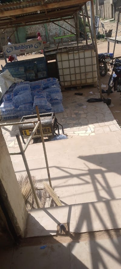 The image depicts an outdoor commercial scene in Danbare, Nigeria, under bright daylight, viewed from a slightly elevated perspective.

The foreground features a tiled pavement area with a black and yellow FIRMAN SPG 2900 FE gasoline generator (2.2kW) positioned on it. Several traditional brooms, made of dried plant material, lean against a metal frame on the left side of the pavement. Strong shadows are cast across the tiled surface, indicating sunlight from the upper right.

Beyond the pavement, a makeshift shelter constructed from wooden posts and corrugated metal roofing covers a significant portion of the scene. Under this shelter, numerous stacks of clear plastic sachets, labeled "HIYCO" (partially visible) and "Habibiyya Water," are piled on wooden pallets. Adjacent to these stacks are two large, white intermediate bulk containers (IBCs) encased in metal cages. A prominent blue and white sign displaying "Habibiyya Water" and a logo stands centrally.

Two individuals are visible further back under the shelter. One individual, wearing light-colored clothing and a cap, is partially obscured by the "Habibiyya Water" sign and holds a red object. Another individual, wearing a pink hijab and dress, walks towards a light green-colored concrete or masonry wall in the background.

To the right of the shelter, two motorcycles are parked on a dirt ground surface. Various debris, including plastic items and dark-colored bags, are scattered across the dirt and tiled areas. The background shows a dirt road and additional simple buildings.