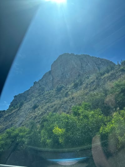 The image shows a view of a hillside from inside a vehicle. A rocky cliff face is visible on the left side of the hillside, while the right side is covered in green vegetation. The sky is blue with the sun shining brightly in the corner, casting a glare. The bottom portion of the image shows a dark, undefined portion of the vehicle's interior. The location of the image is in el Verger, Spain.