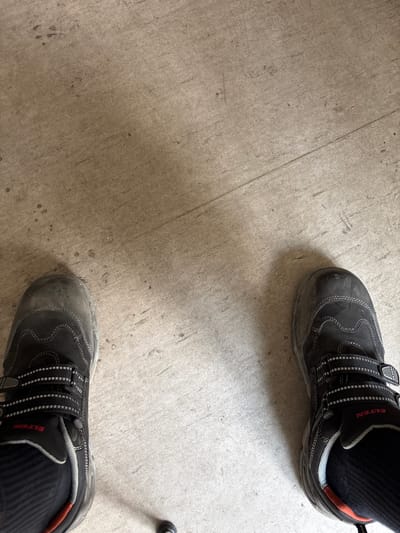 The image shows a top-down view of two black work boots on a light gray concrete floor. The boots are positioned diagonally across the frame, with one boot in the upper right corner and the other in the lower right corner. Both boots appear to be worn and slightly dusty. They are designed for work, with reinforced toes and sturdy soles. Each boot has multiple straps with Velcro fasteners for adjustment, and reflective stitching. The brand name "ELTEN" is visible in red text on the side of the boot in the upper right. The concrete floor shows some scuff marks and variations in tone, suggesting an indoor industrial or workshop setting. The lighting appears to be artificial and even, indicating the image was taken indoors. No people are visible, only their feet wearing the boots. The overall impression is utilitarian and functional, suggesting the wearer is engaged in some form of manual labor or work. The scene is devoid of any clear indicators of time of day or weather, and the lack of surrounding context makes the specific location difficult to determine beyond it being a concrete floor.