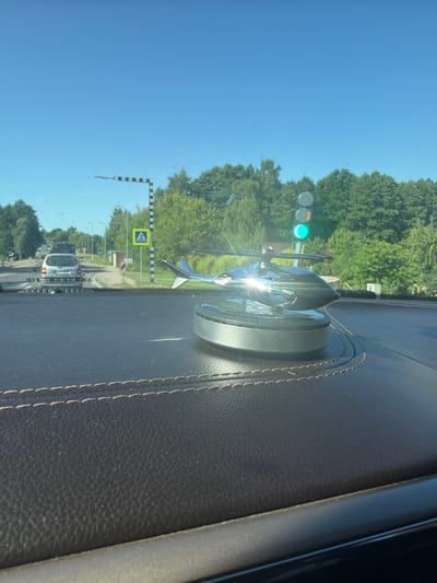 A close-up, eye-level shot shows a shiny, metallic helicopter figurine on a dark, textured dashboard inside a vehicle. The helicopter is positioned in the foreground to the right of the image, reflecting sunlight. Beyond the dashboard, through the windshield, a street scene unfolds under a clear blue sky. A white car is visible on the road, followed by a dark-colored vehicle. Traffic lights, pedestrian crossing signs, and trees line the street. The environment suggests a sunny day in an urban or suburban area, likely Vecstropi, Latvia, given the presence of a pedestrian crossing sign and the style of road infrastructure. There are no people visible in the frame.