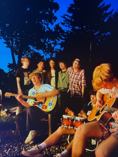 A group of young people are gathered outdoors, likely in a backyard or park setting, as indicated by the silhouette of trees and a clear blue sky. The image appears to be taken at dusk or early evening, with the blue sky suggesting daylight is fading. The people are casually dressed, with one individual playing an acoustic guitar decorated with stickers. Another person is seated next to a snare drum. The overall atmosphere suggests a relaxed, informal gathering, possibly a jam session or a small outdoor concert. Some individuals are looking at the guitar player, while others are engaged in conversation or observing the scene. The mood seems to be one of enjoyment and camaraderie. There is no visible text in the image.