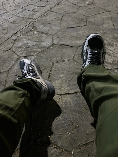 The image shows a person's legs and feet wearing olive green pants and grey and black athletic shoes. The shoes have a chunky sole and intricate detailing. They are standing on a textured, grey stone surface that appears to be paving stones arranged in an irregular pattern. The lighting suggests it is either nighttime or a dimly lit indoor space, casting shadows from the person's legs onto the ground. The environment is consistent with an outdoor urban setting, possibly a public walkway or plaza. There is no indication of the specific location being in Tipaza, Algeria.
