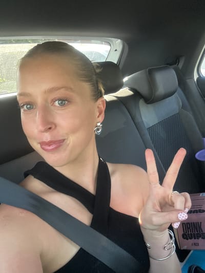 A woman is sitting in a car wearing a black top, a seatbelt, and several silver bracelets. Her nails are painted light pink with red polka dots. She is holding up two fingers in a peace sign. A purple bottle and a box of "DRINK QUIPS" are in the background. The location is possibly Thirroul, Australia, based on the prompt.