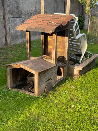The image shows an outdoor scene in Jelgava, Latvia, bathed in the warm light of what appears to be late afternoon or early evening. The primary subject is a weathered wooden structure, possibly a playhouse or an abandoned shed, situated on a grassy area. This structure has a pitched roof covered with what looks like rusted asphalt shingles. It features open sections, revealing the interior which appears to be empty or filled with debris. A small, rusty metal wheel is visible attached to the side of the structure.
Adjacent to the wooden structure, a white plastic chair is placed on top of other wooden elements, possibly part of a disassembled structure or debris. The background consists of more grass, a concrete wall or fence in the lower left, and a chain-link fence with vegetation behind it. The overall impression is one of disuse and perhaps neglect, with the natural elements of grass and greenery encroaching on the man-made objects. There is no visible text in the image. The lighting suggests a calm, possibly summer evening.