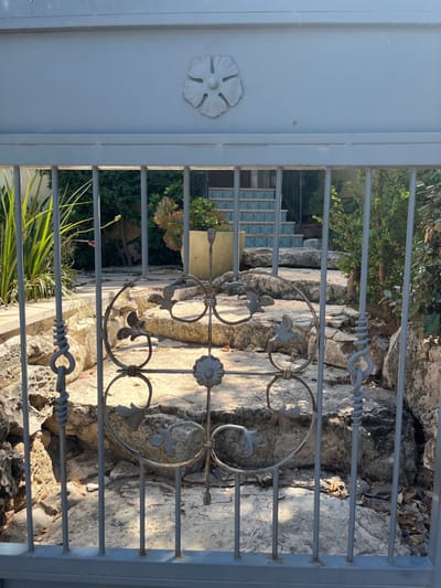 The image depicts a head-on view through a closed, grey metal gate into a residential property located within the Menashe Regional Council, Israel. The gate features a prominent horizontal top rail adorned with a central, raised, light-colored floral or stylized leaf motif. Below this, vertical grey bars descend, with a decorative wrought-iron section integrated into the lower center, displaying ornate scrolls, stylized leaf elements, and a central rosette design.
Beyond the gate, an outdoor, sunlit environment is visible. An uneven path composed of large, irregularly shaped natural stone steps ascends into the property from the foreground towards the background. To the left of this path, tall, green, blade-like plants are present. In the mid-ground, a light-colored, possibly beige or yellow, planter pot contains a large succulent plant with reddish-green foliage. Further into the property, a more formal set of steps is visible, with risers adorned in blue and white patterned tiles. Lush green vegetation, including various trees and shrubs, flanks both sides of the ascending path and fills the background. Sunlight casts defined shadows across the scene, indicating clear weather. No individuals are present in the image.