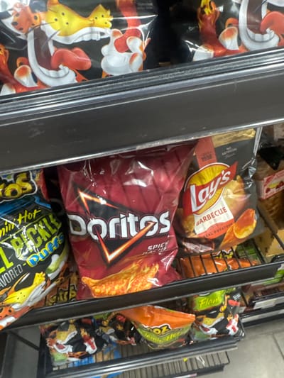 In Lake City, United States, this image captures a close-up view of snack items displayed on metal shelves. The focus is on bags of chips, with a notable presence of Doritos, Lay's Barbecue, and what appears to be Cheetos with a character illustration. The items are packaged in colorful bags, with some showing visible texture of the chips inside. The shelves are dark gray and show some wear. The lighting appears to be artificial, typical of an indoor retail environment. The perspective is from slightly above, looking down at the products.