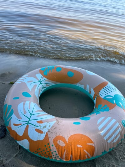 An inflatable swim ring with a tropical leaf and polka dot pattern rests on the sandy shore of a body of water. The swim ring is predominantly beige with accents of orange, teal, and white. Small water droplets are visible on its surface. The water behind the swim ring is calm with gentle ripples and reflects the sky. The sand appears damp, suggesting the proximity of the water. The scene is set outdoors, likely at a beach or riverbank, with no people visible. The location is identified as Lyutizh, Ukraine.