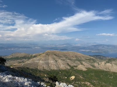 This is a scenic landscape photograph taken from a high vantage point, likely a mountain, looking out over rolling hills, a body of water, and distant mountains under a blue sky with scattered clouds.

The foreground features rocky, grey terrain with sparse green vegetation. A dark green tree is visible on the left. The mid-ground is dominated by undulating, tan-colored hills with a winding white road snaking its way up one of the slopes. Patches of darker green vegetation are scattered across the hillsides.

In the background, a calm, dark blue body of water, likely a sea or large lake, stretches out towards a coastline dotted with buildings. Further in the distance, a range of hazy, blue-grey mountains forms the horizon. The sky is a bright blue, with dynamic white clouds, some appearing wispy and others more substantial, suggesting a pleasant, partly cloudy day. The lighting indicates it is daytime, likely late morning or early afternoon, given the brightness and the shadows cast.

The scene evokes a sense of natural beauty and tranquility, with the winding road suggesting human presence and accessibility to this remote location within the Kassopaia Municipal Unit, Greece. There is no visible text in the image.