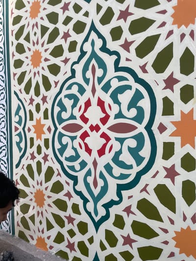 An ornate painted ceiling displays Islamic-inspired geometric patterns. A central arabesque design features red, teal, and brown hues, surrounded by a creamy white framework. Around it, a network of geometric stars and polygons utilizes shades of orange, green, and dark red. A border at the bottom shows a repetitive, stylized leaf pattern, painted in white. The overall effect is rich in detail and color. A section of a person is visible at the bottom of the image.