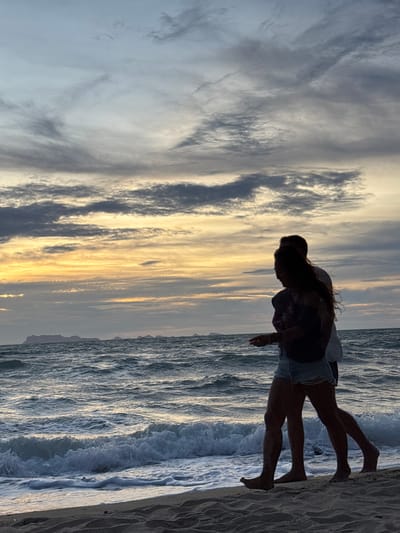 A couple walks along a sandy beach in Tong Yang, Thailand, at sunset. The sky is a dramatic display of clouds in various shades of gray, blue, and orange, with the sun setting on the horizon, casting a warm glow over the scene. The ocean waves crash gently onto the shore, creating a foamy white line of surf. The silhouettes of the couple are visible against the vibrant sky, suggesting an intimate and romantic moment. The man's arm is around the woman as they walk side-by-side, their legs and feet appearing in the foreground. The sand is damp from the receding tide, with footprints visible. In the distance, several islands are faintly visible on the horizon, adding to the picturesque landscape of Tong Yang.