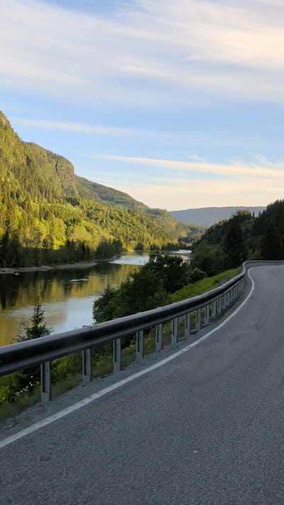 The image depicts a scenic view of the Namsen river flowing through a valley in Grong, Norway. The road, with a metal guardrail, curves to the right in the foreground, suggesting a driver's perspective. Lush green trees cover the steep slopes of the valley on both sides, reflecting in the calm water of the river. The sky is a clear blue with scattered white clouds, and the sunlight casts long shadows, indicating it might be late afternoon or early morning. The overall impression is one of natural beauty and tranquility.