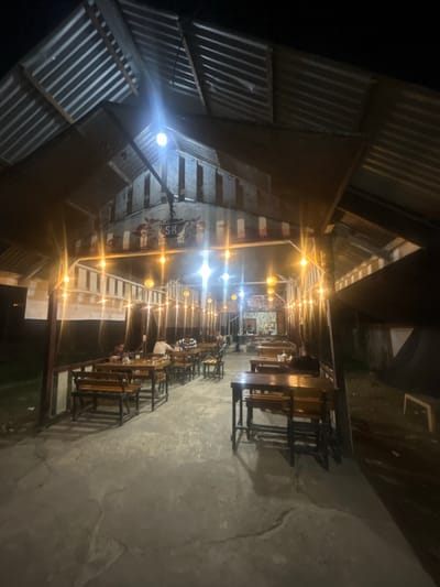 This image depicts an outdoor seating area of a restaurant or cafe at night. The location is identified as Keudematangkuli, Indonesia. The scene is illuminated by numerous string lights and overhead lights, casting a warm glow on the wooden tables and benches arranged on a concrete floor. The structure appears to have a corrugated metal roof supported by wooden beams. In the background, there is a mural or sign with the text "SK Semu Kupie" and a stylized coffee cup. Several patrons are visible seated at the tables, engaged in dining or conversation. One person is seen walking away from the camera towards the back of the establishment. The overall atmosphere is casual and inviting, typical of a nighttime eatery.